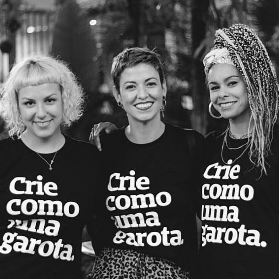 Foto preta e branca com 3 mulheres. Da direita para a esquerda: mulher negra com tranças escuras e claras no cabelo, presas em um rabo. No meio, mulher branca com cabelos curtos, lisos e escuros e a da esquerda, mulher branca com cabelo chanel, platinado. Todas estão sorrindo na foto e abraçadas. Elas vestem peita preta com a frase crie como uma garota em branco. Fundo desfocado.
