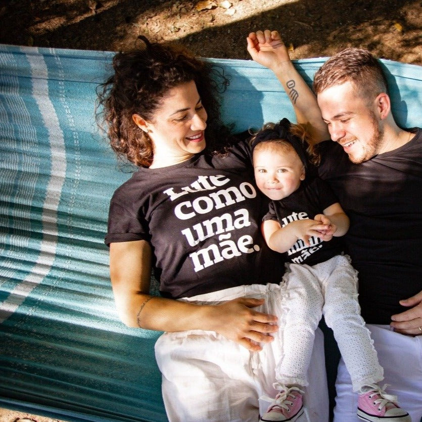 Foto colorida de uma família deitados em uma rede, a mãe mulher branca de cabelos cacheados escuros veste uma camiseta rpeta com frase Lute como uma mão em branco, a filha no meio do casal criança branca de cabelos claros veste peitinha preta com frase Lute como minha mãe em brando, usa faixa preta na cabeça e morde lábio inferior e sorri. Pai homem branco de cabelos curtos e claros, veste camiseta preta lisa. O casal olha e sorri para a criança no meio. todos de vestem calça branca.