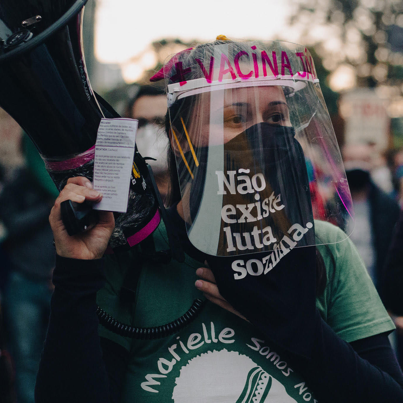 Foto colorida de uma mulher branca de cabelos castanhos e presos, usa um lenço de guerrilha preto escrito Não existe luta sozinha em branco, está com uma máscara de proteção facial transparente e segura um megafone