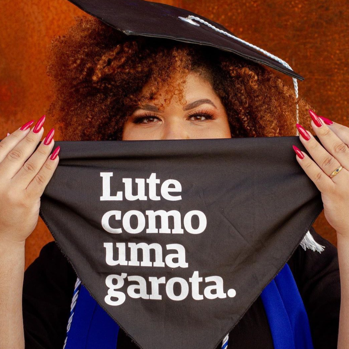 Foto colorida de uma mulher negra de cabelos crespos, black power, soltos, ruivos e segura um lenço preto escrito lute como uma garota em branco. Ela está vestida com uma beca de formatura.