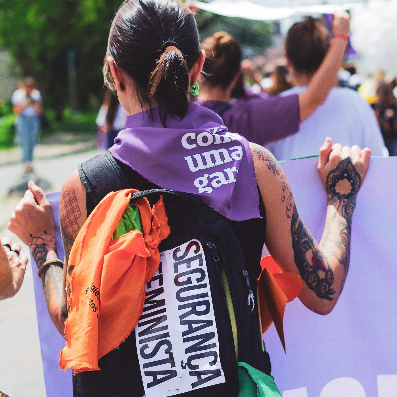 Foto colorida de uma mulher branca de cabelos castanhos, lisos, presos em rabo, e está com lenço roxo com a frase lute como uma garota em branco amarrado no pescoço. Está de costas, com uma mochila preta com um adesivo escrito segurança feminista.