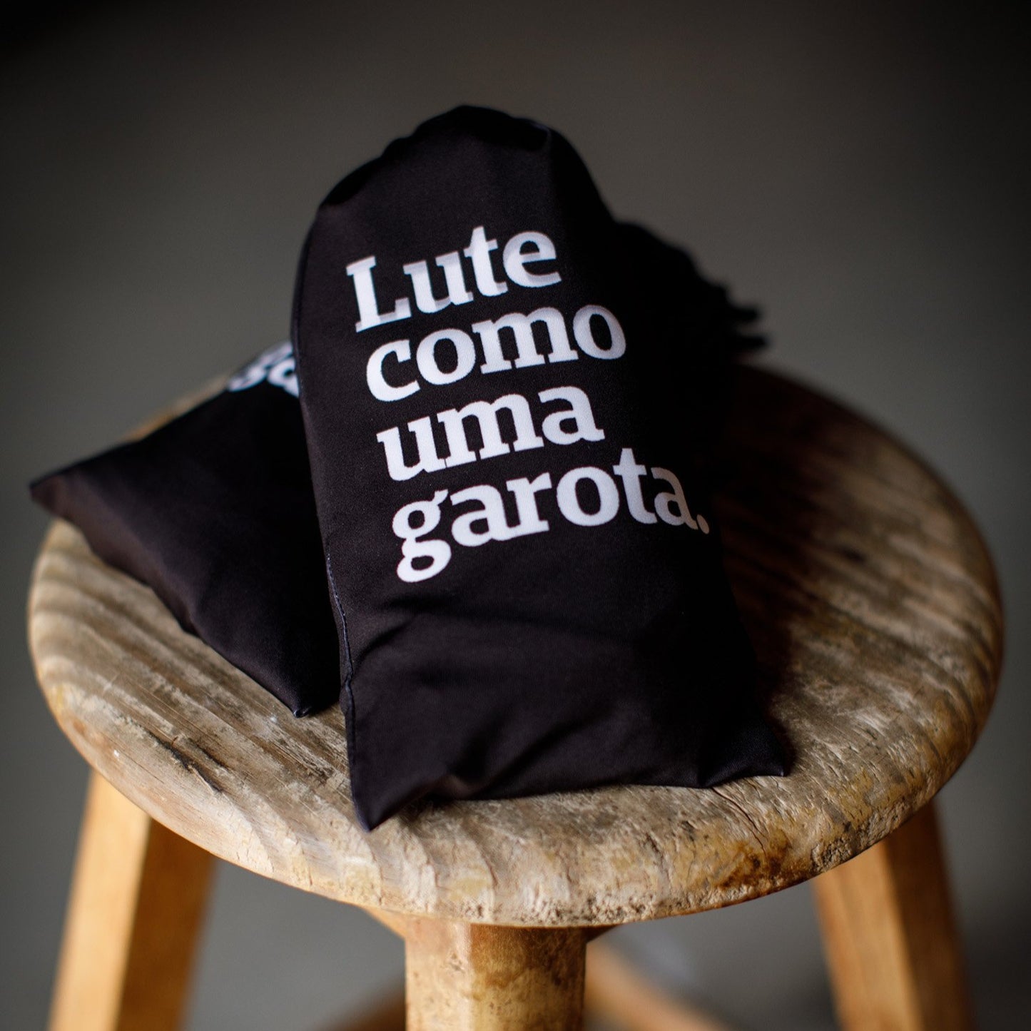 Foto colorida de dois sacos de rash guard preto escrito Lute como uma garota em branco, ambos estão sob uma banquetinha redonda de madeira cru.