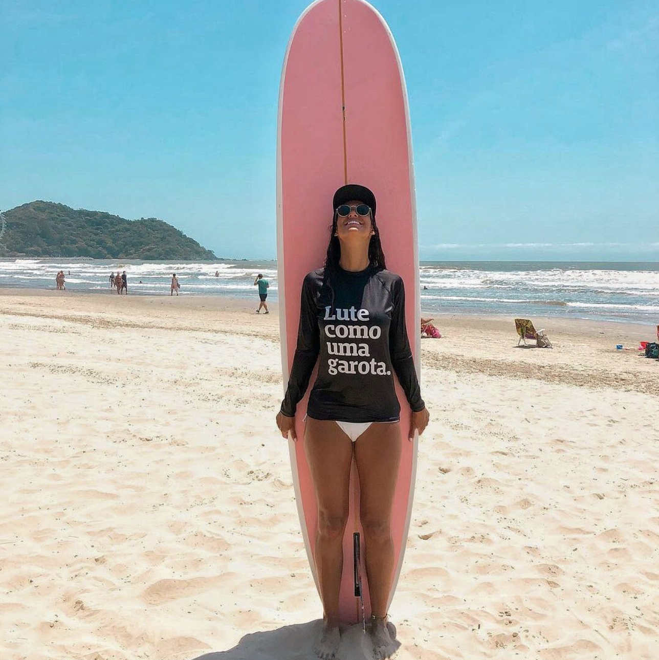 Foto colorida de mulher branca de cabelos escuros molhados e soltos, veste uma rash guard de lycra com a frase Lute como uma garota em branco e calcinha de biquini branca, está na frente de uma prancha se surfe rosa, com a cabeça erguida, usa boné preto e óculos de sol, ao fundo praia.