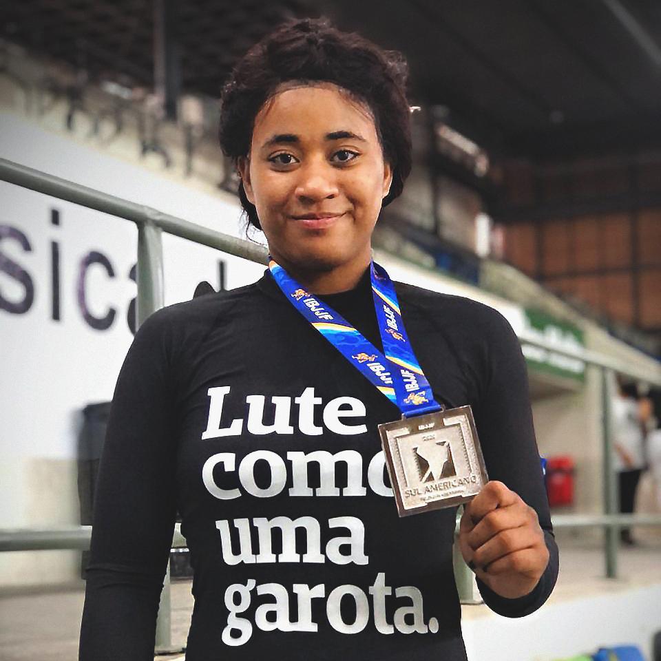 Foto colorida de uma mulher negra, cabelos castanhos, presos, com rash guard de manga comprida, preta, escrita luta como uma garota em branco. Ela tem uma medalha prata no pescoço e a segura com a mão direita. Sorri na foto.