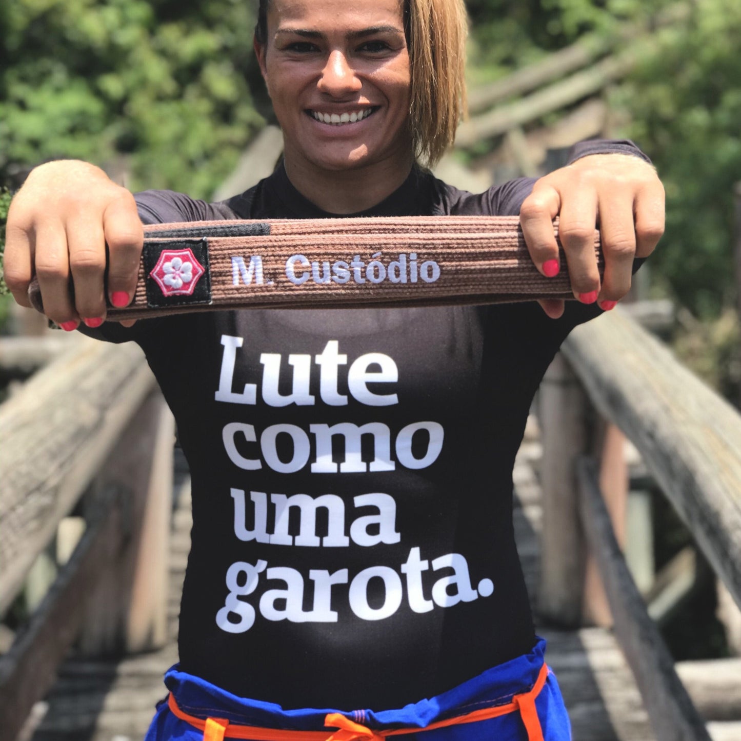 Foto colorida de uma mulher branca, cabelos castanhos, mechas claras, preso em rabo, vestida com rash guard de manga comprida, preta, escrita lute como uma garota em branco. Segura nas mãos mãos uma faixa marrom escrito M Custódio em branco, está sorrindo pra foto.