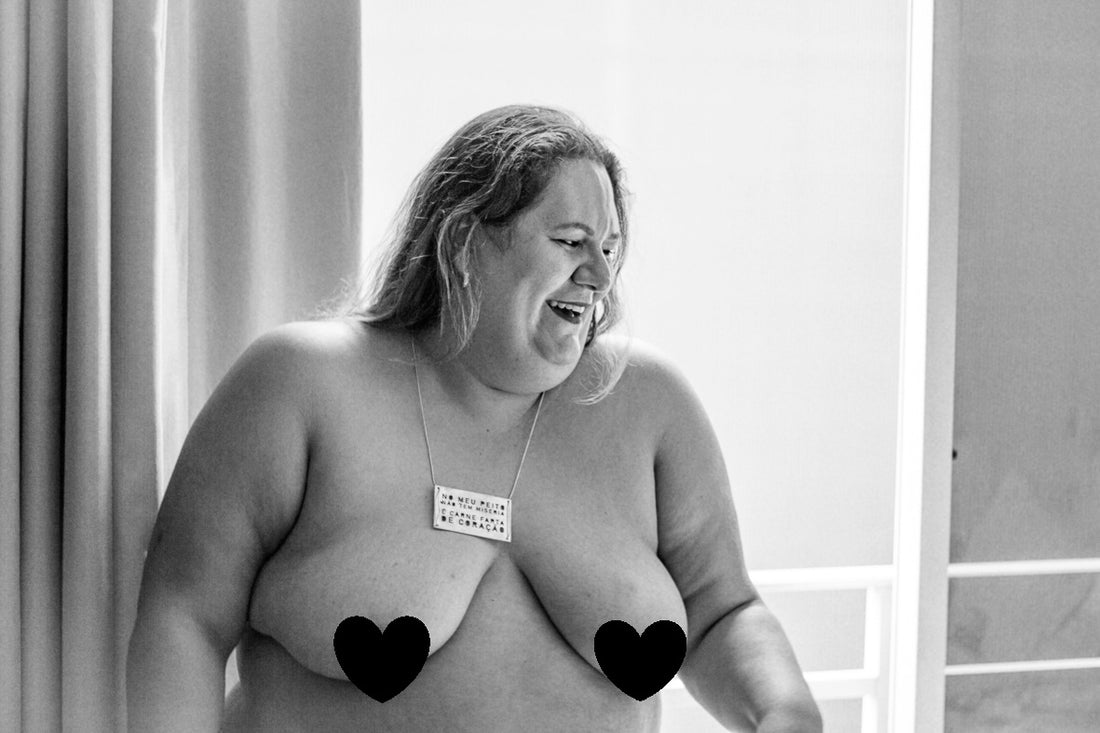 foto preto e branca da jéssica, mulher branca, com cabelos castanhos claros, lisos, compridos, com um colar com pingente retangular, parada em frente a uma sacada, com mamilos cobertos com coração