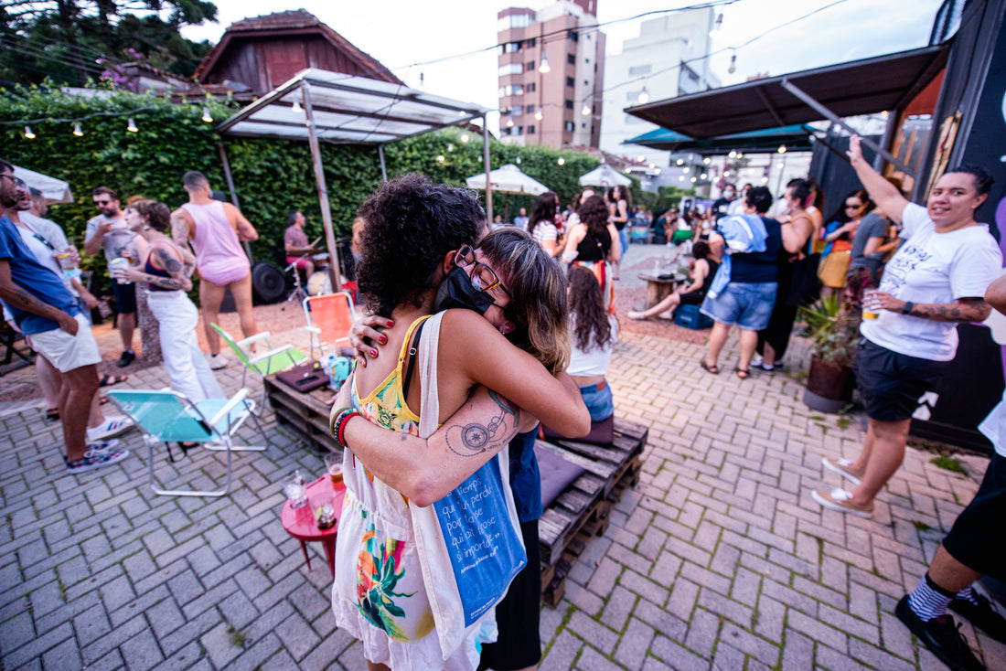 foto colorida em ambiente externo de duas mulheres se abraçando e ao fundo, pessoas em pé, sentadas, bebendo chopp. Também tem containers, guarda-sol e mesas com cadeiras.