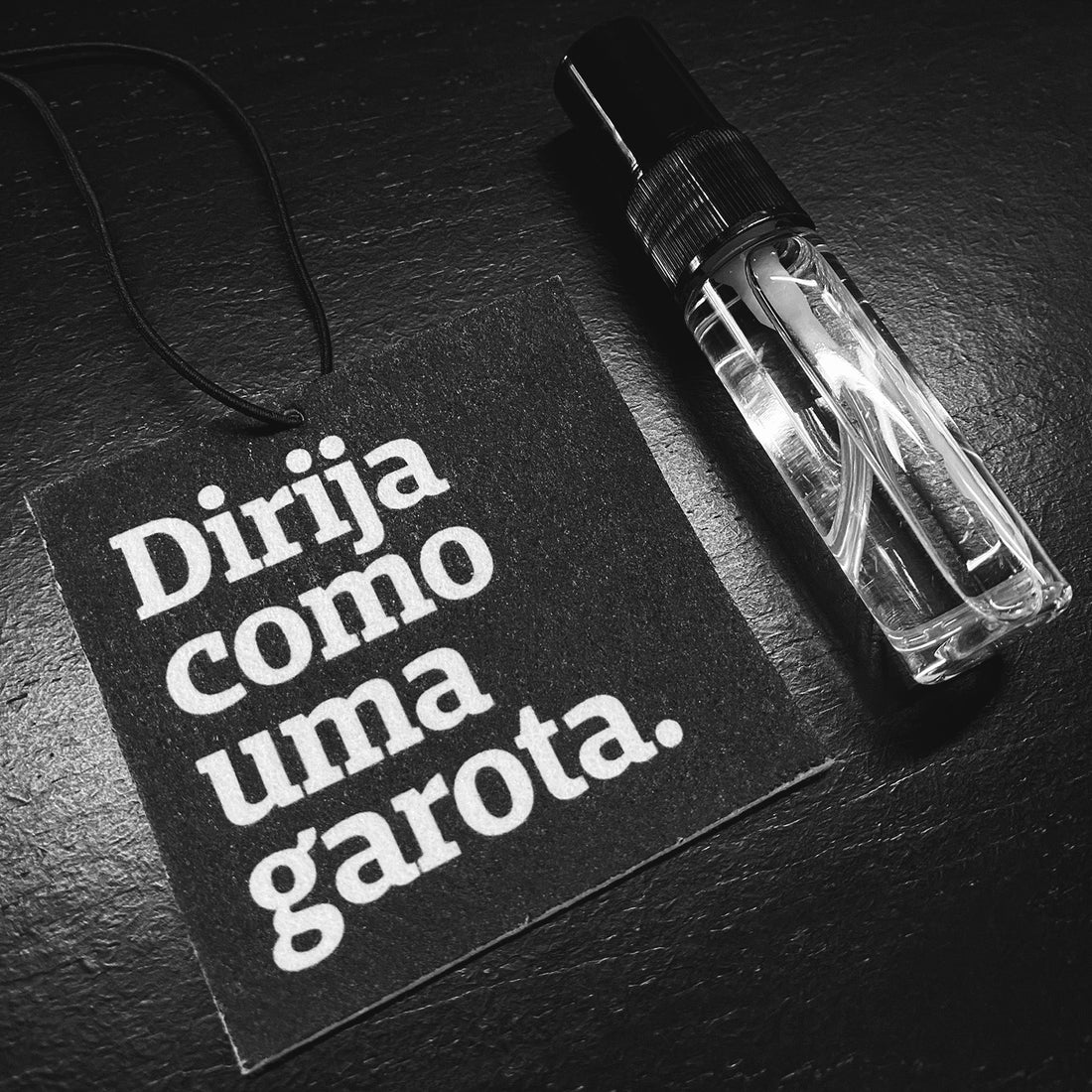 foto preto e branca de um aromatizador automotivo quadrado, preto, com a frase dirija como uma garota em branco e uma alça preta. Tem também um frasco transparente com líquido dentro, disposto em cima de uma mesa preta.