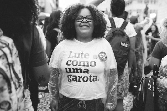 mulher negra com cabelos encaracolados na altura do ombro. esta em meio a uma manifestação, parada, sorrindo para a câmera. Ela veste uma peita branca com a frase 'Lute como uma garota.' em preto e óculos com armação preta. 
