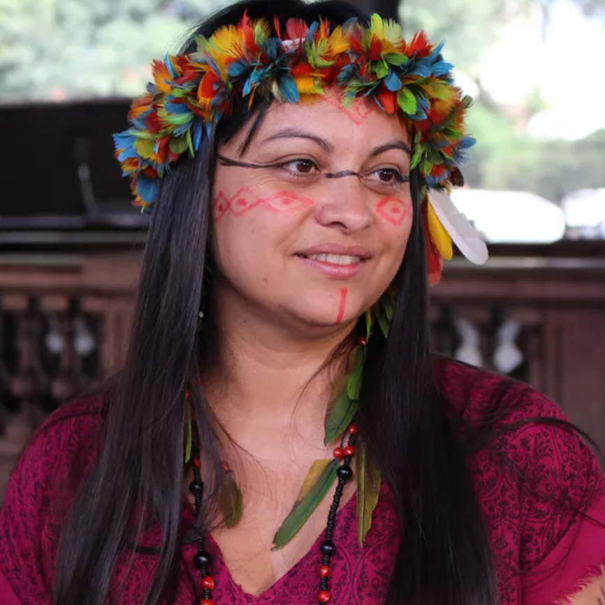 foto colorida da Fabiane Cruz, mulher, indígena, cabelos compridos, lisos, pretos, com cokar de penas pequenas e coloridas ao entorno da cabeça, pintura nos olhos, bochechas e queixo. está com uma blusa bordô e usa brincos de pena e miçanga
