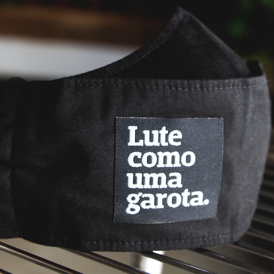 foto preto e branco do rosto de uma mulher, negra, cabelos trançados, castanhos, compridos. Está com uma máscara preta com um patch preto com a frase seja quem você quiser em branco. 