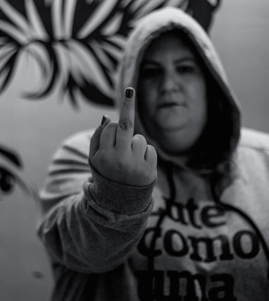 foto preto e branca da Jéssica, mulher branca, vestida com um moletom cinza e a frase lute como uma garota em preto. Ela está de capuz, mostrando o dedo do meio. 