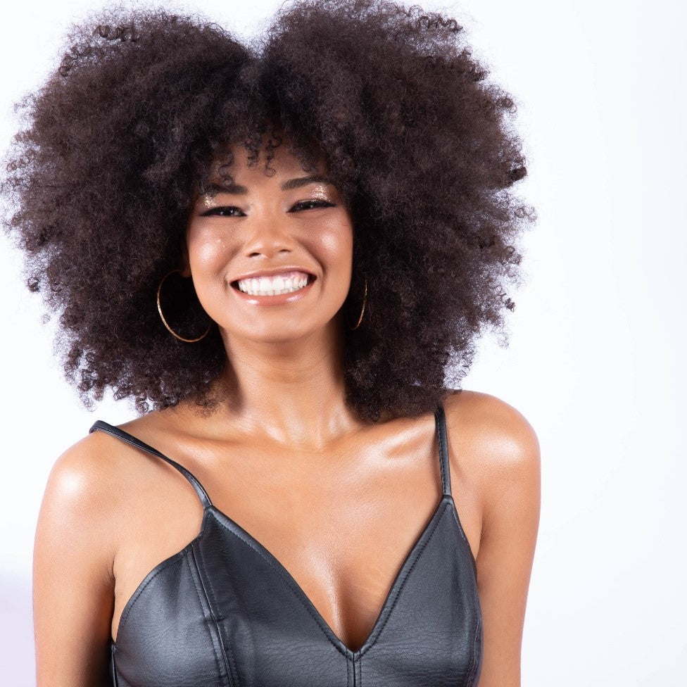 foto colorida da Amanda, mulher negra, com cabelos castanhos, crespos e médios. Está usando batom vermelho e camisa com flores amarelas e folhas azuis. Está com o indicador esquerdo no queixo e sorri pra foto. 