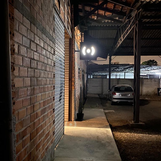 foto do letreiro da peita, um quadrado preto com a logo, um peitinho, ( . ), em branco. O luminoso está aceso. A parede da lateria esquerda é de tijolos e a porta de entrada é de ferro. 