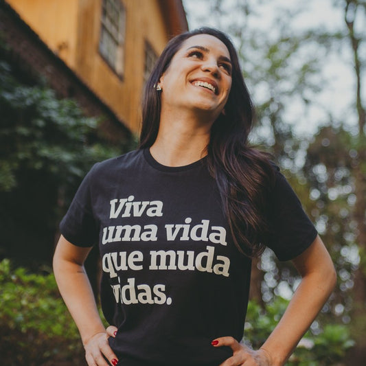 Foto colorida de uma mulher branca de cabelos castanho lisos, longos e soltos, com as mãos na cintura, olha para cima e sorri. Veste camiseta preta com a frase Viva uma vida que muda vidas em rbanco. Fundo de folhagens desfocadas