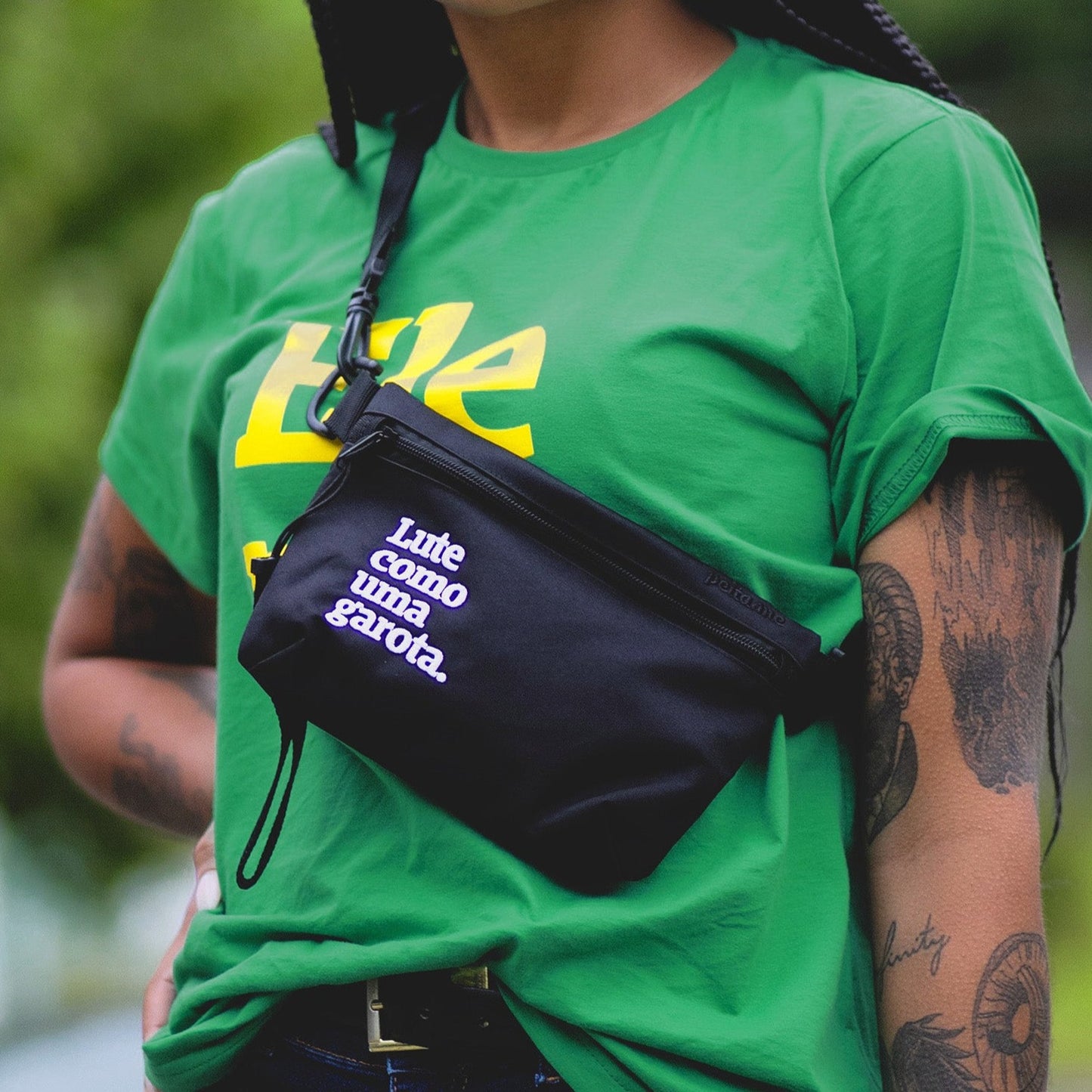 Foto colorida de uma mulher negra de cabelos compridos, castanhos e trançados com uma pochete preta escrito lute como uma garota em branco, está posando, em ambiente externo, com árvores desfocadas ao fundo, Ela veste uma camiseta verde escrito ele não em amarelo.