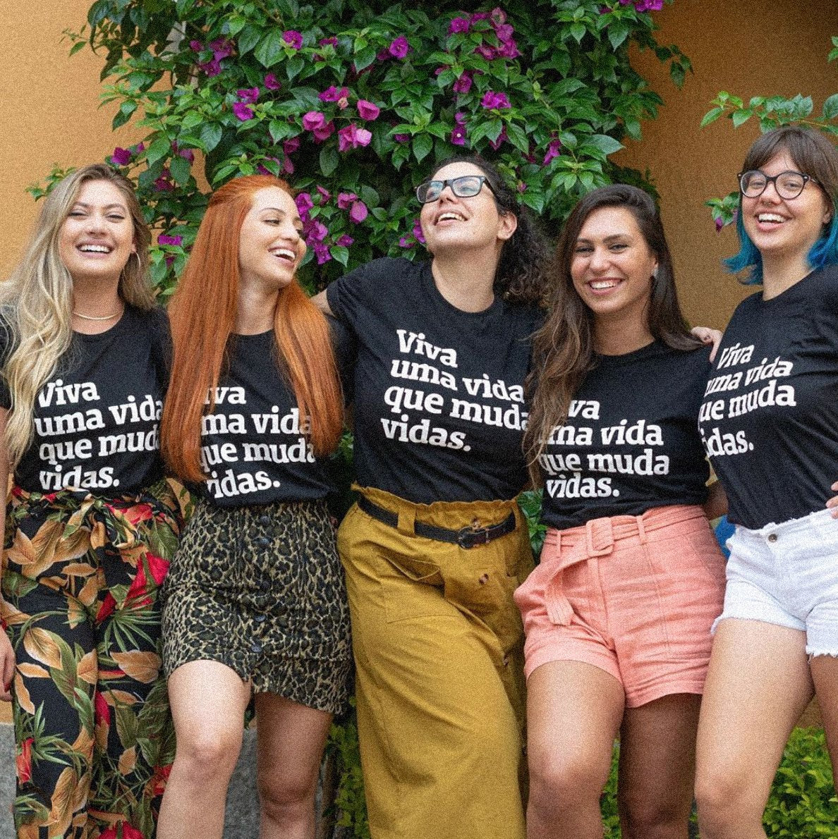 Foto colorida de 5 cinco mulheres brancas, da esquerda para direita,mulher 1: cabelos loiros, longos, lisos, soltos, brincos e colar prateado. Mulher 2: cabelos ruivos, lisos, longos, soltos. Mulher 3: cabelos castanhos, enrolados, chanel, meio preso, usa óculos de grau. Mulher 4: cabelos castanhos, lisos, longos, soltos. Mulher 5: cabelos castanhos e azul nas pontas, lisos, chanel, tem franja e usa óculos de grau. Todas vestem camiseta preta escrita viva uma vida que muda vidas em branca e sorriem na foto.