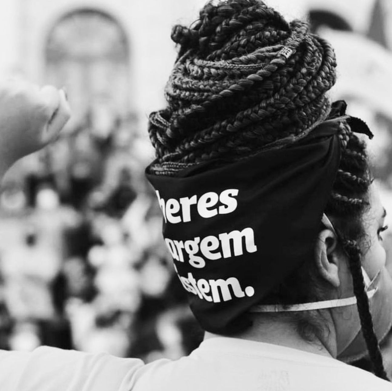 Foto preto e branco da Amanda, mulher negra, com cabelos trançados, preso em coque, com lenço de guerrilha preto escrito mulheres a margem resistem em branco amarrado na cabeça.