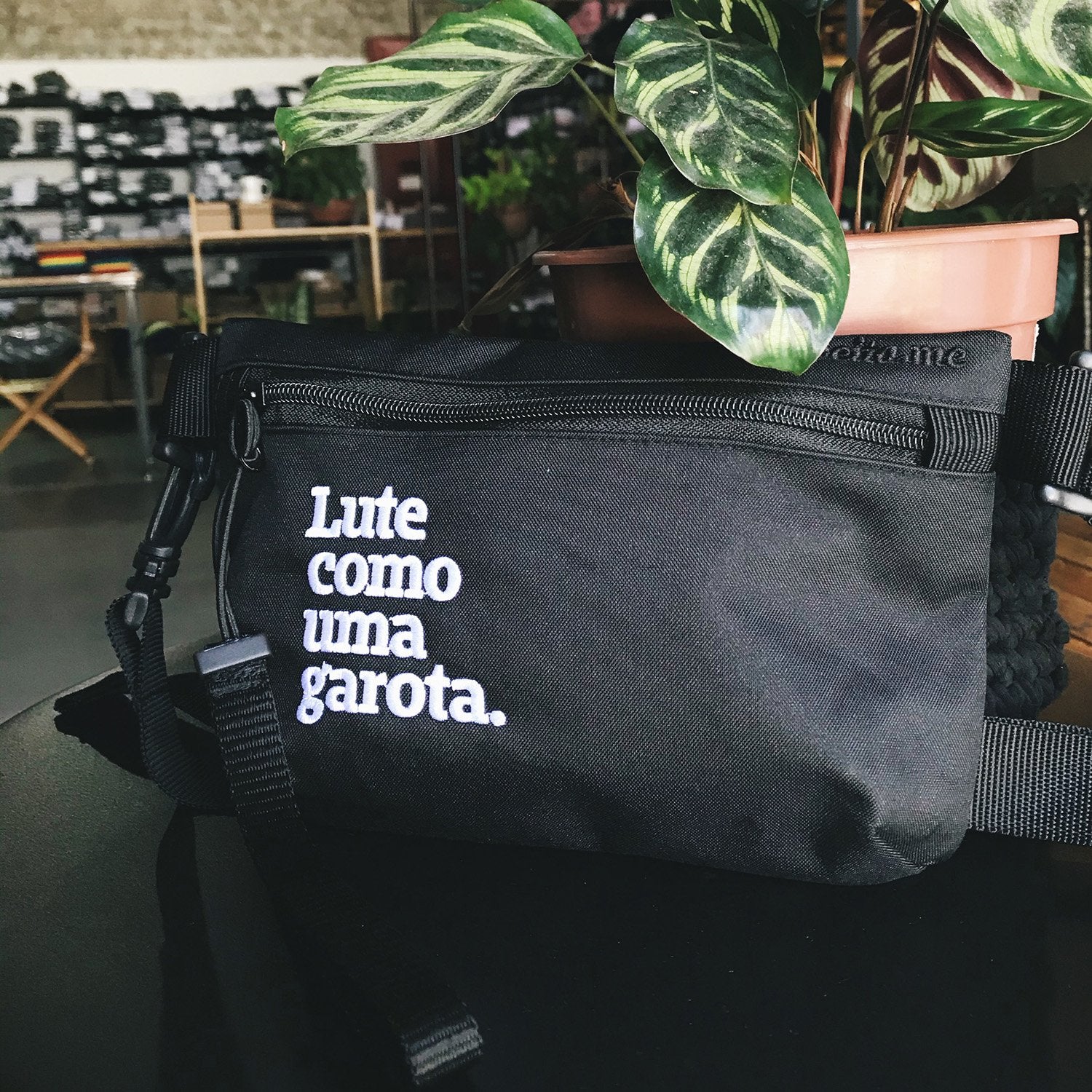 Foto colorida de uma pochete preta escrito lute como uma garota em branco. Ela está encostada em um vaso de planta em cima de uma mesa preta.