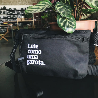 Foto colorida de uma pochete preta escrito lute como uma garota em branco. Ela está encostada em um vaso de planta em cima de uma mesa preta.