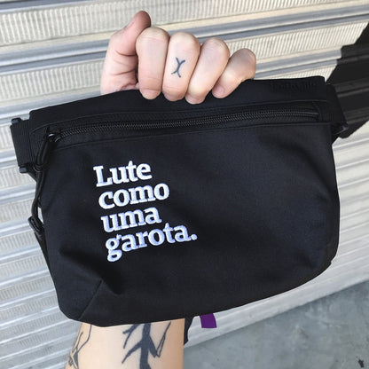 Foto colorida de uma mão branca segurando uma pochete preta escrita lute como uma garota em branco.