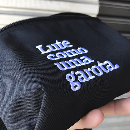 Foto colorida de uma mão branca segurando uma pochete preta com foco na frase lute como uma garota em branco.