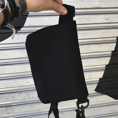 Foto colorida de uma mão branca, com pulseiras, segurando a alça de uma pochete preta, mostrando a parte de trás.