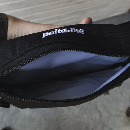 Foto colorida de uma mão branca mostrando a parte interna de pochete preta, por dentro branca, em cima escrita peita.me, ela mostra o lado de dentro na cor branca.