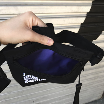 Foto colorida de uma mão branca segurando uma  uma pochete preta aberta, com escrita lute como uma garota em branco seu interior é roxo.